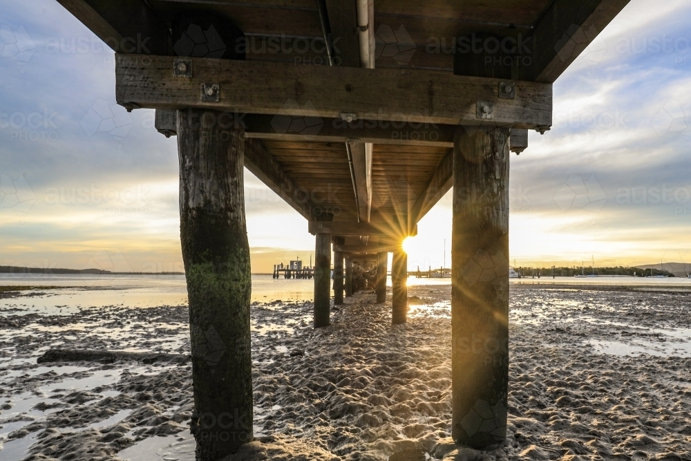 Low angle view of timber jetty leading to sunset through pylons - Australian Stock Image