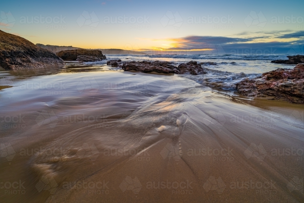 Low angle view of water running off a sandy beach and through a rockpool just before sunrise - Australian Stock Image