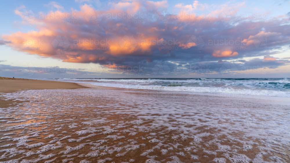 Low angled view of a colourful storm clouds over waves breaking in a sandy beach at twilight - Australian Stock Image