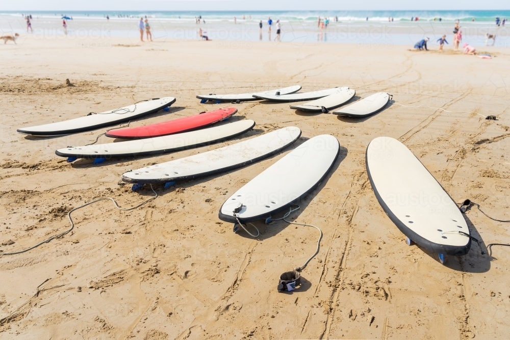 Low angled view of surfboards lying on a sandy beach with beach goers in the background - Australian Stock Image