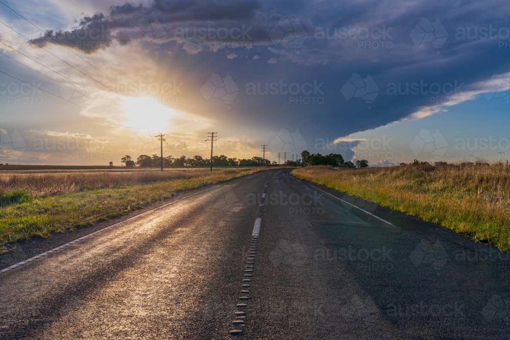 Low angled view of the Sun appearing below a dramatic storm cloud over a country road - Australian Stock Image