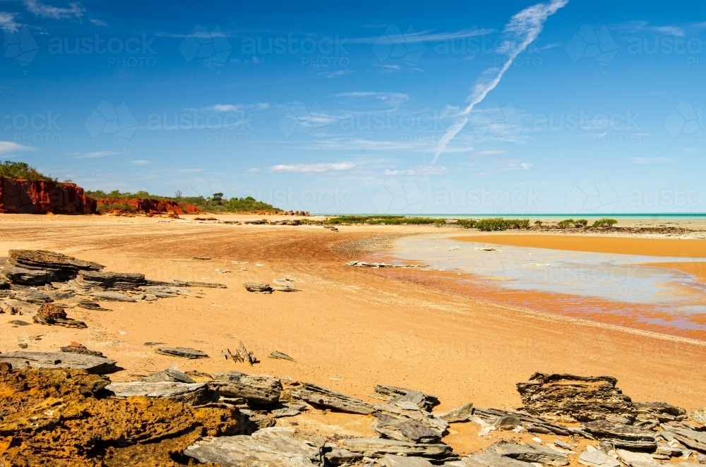 Low tide on an orange sandy beach with red cliffs, rocky foreground and blue sky - Australian Stock Image