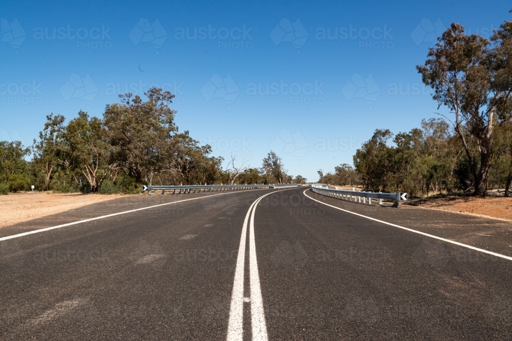 Low view of bitumen road and double white lines leading to horizon.  Trees and clear blue sky - Australian Stock Image