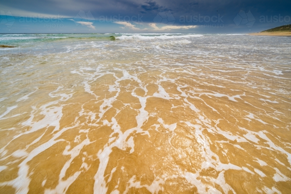 Low wide angle view of waves rolling up a golden sandy beach with dark storm clouds overhead - Australian Stock Image