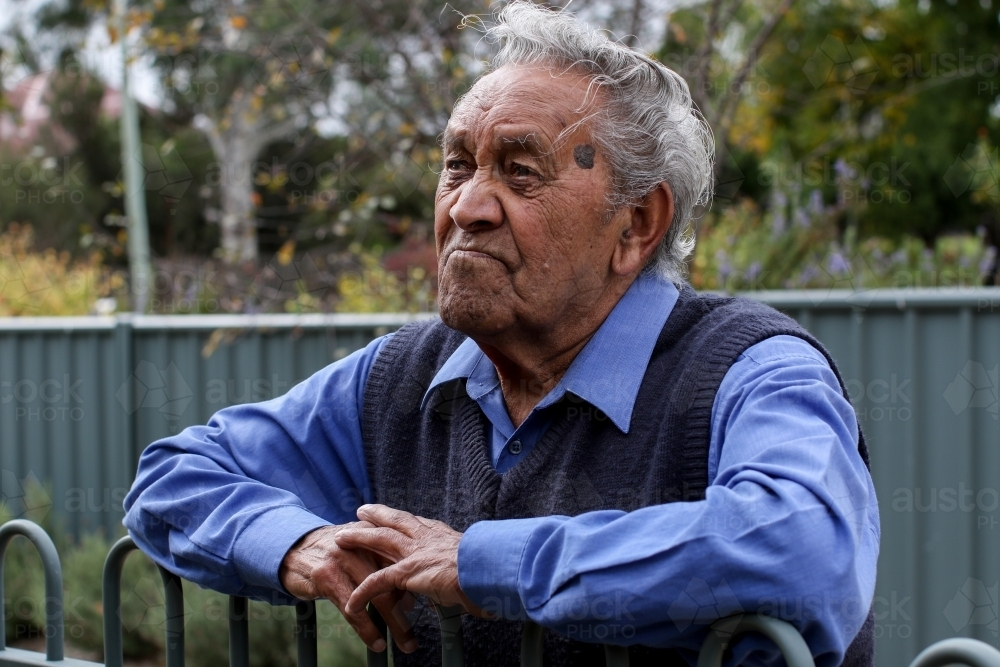 Male Aboriginal elder leaning against fence looking away - Australian Stock Image
