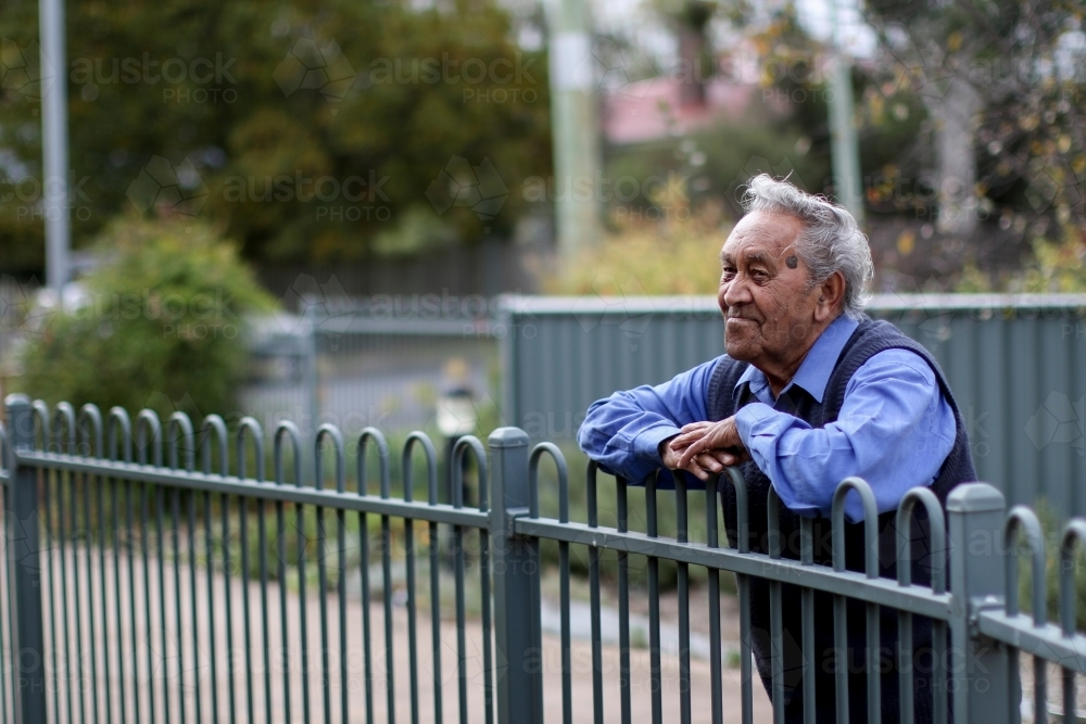 Male Aboriginal elder leaning against fence looking away - Australian Stock Image