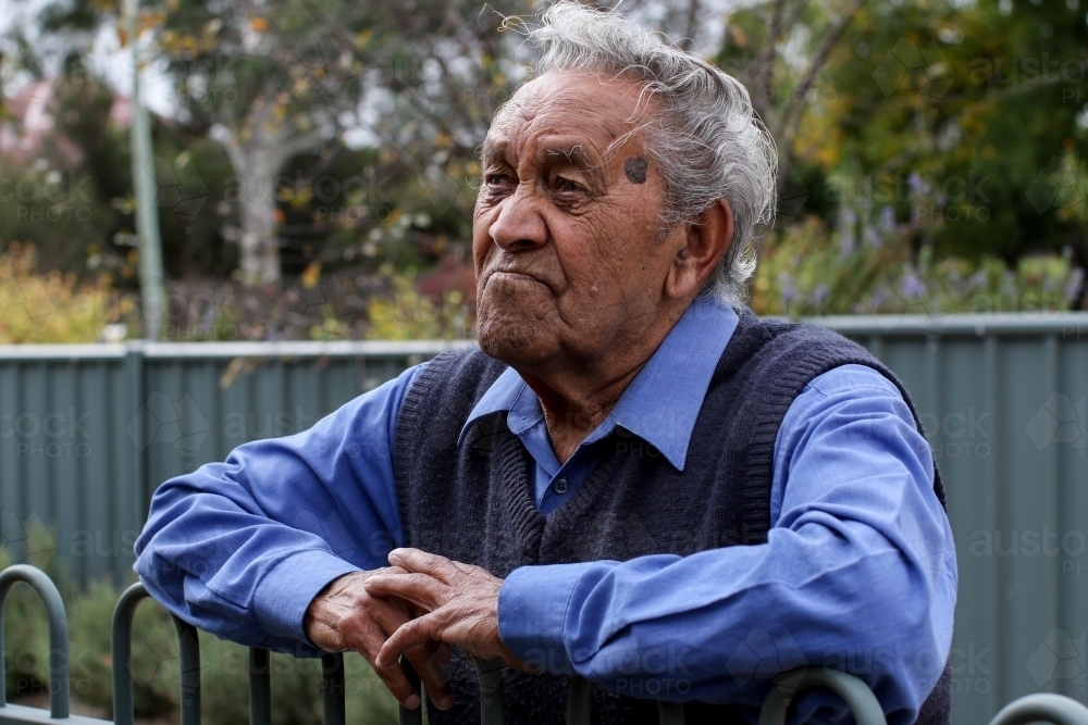 Male Aboriginal elder leaning against fence looking away from camera - Australian Stock Image
