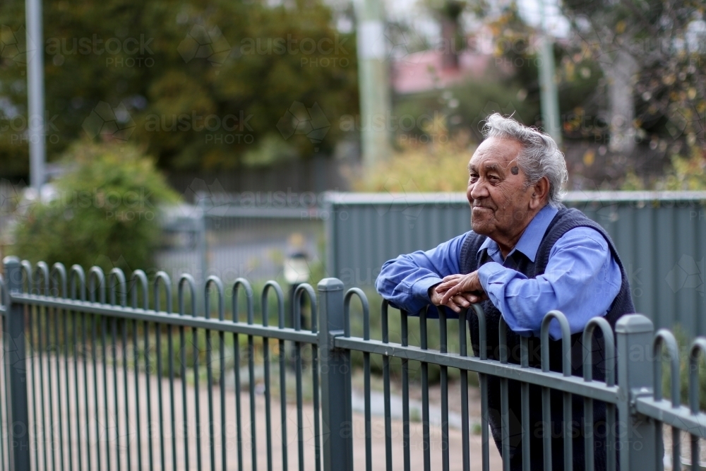 Male Aboriginal elder leaning against fence looking away from camera - Australian Stock Image