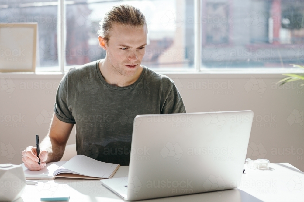 Male designer looking at a laptop screen in a bright white studio - Australian Stock Image