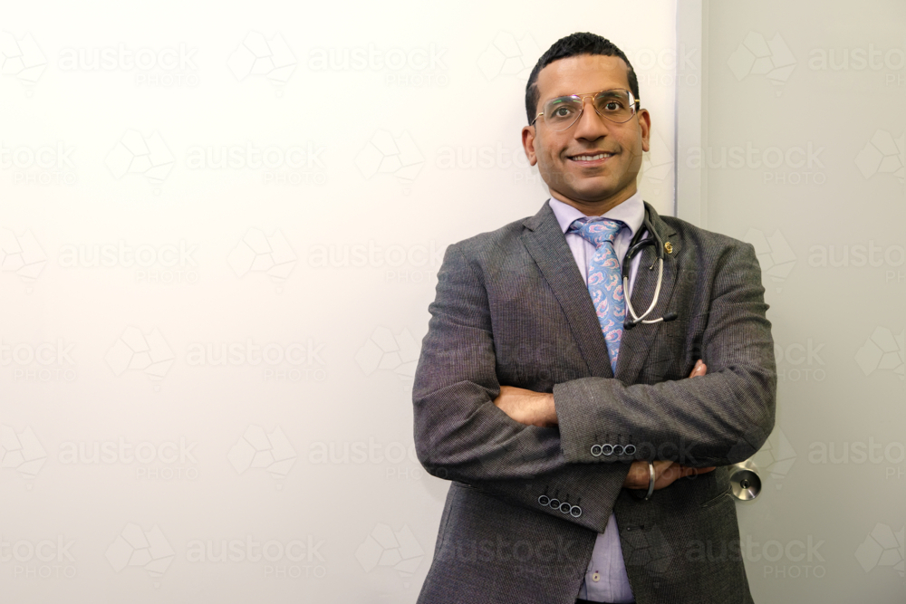 Male doctor standing with arms crossed against white wall - Australian Stock Image