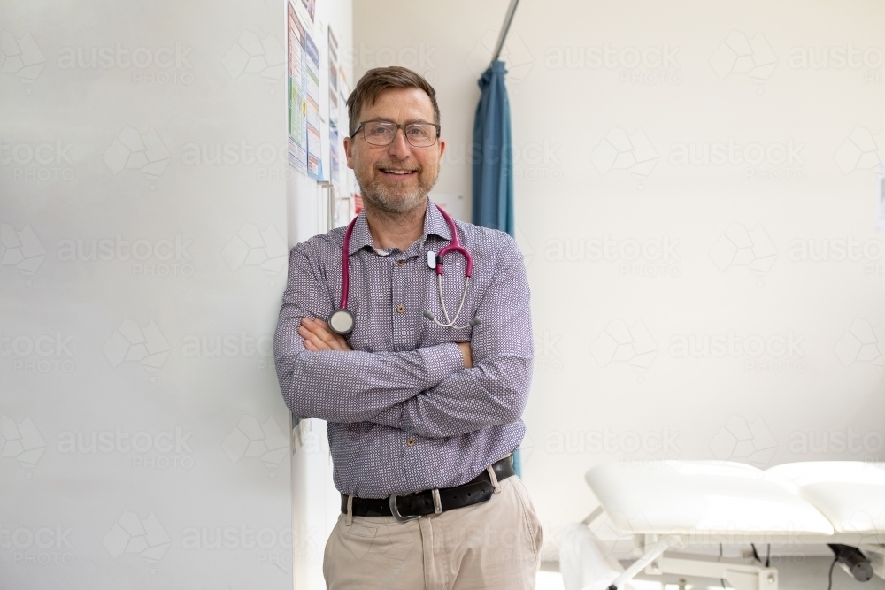 Male doctor with stethoscope around his neck standing while leaning on the wall in the clinic - Australian Stock Image