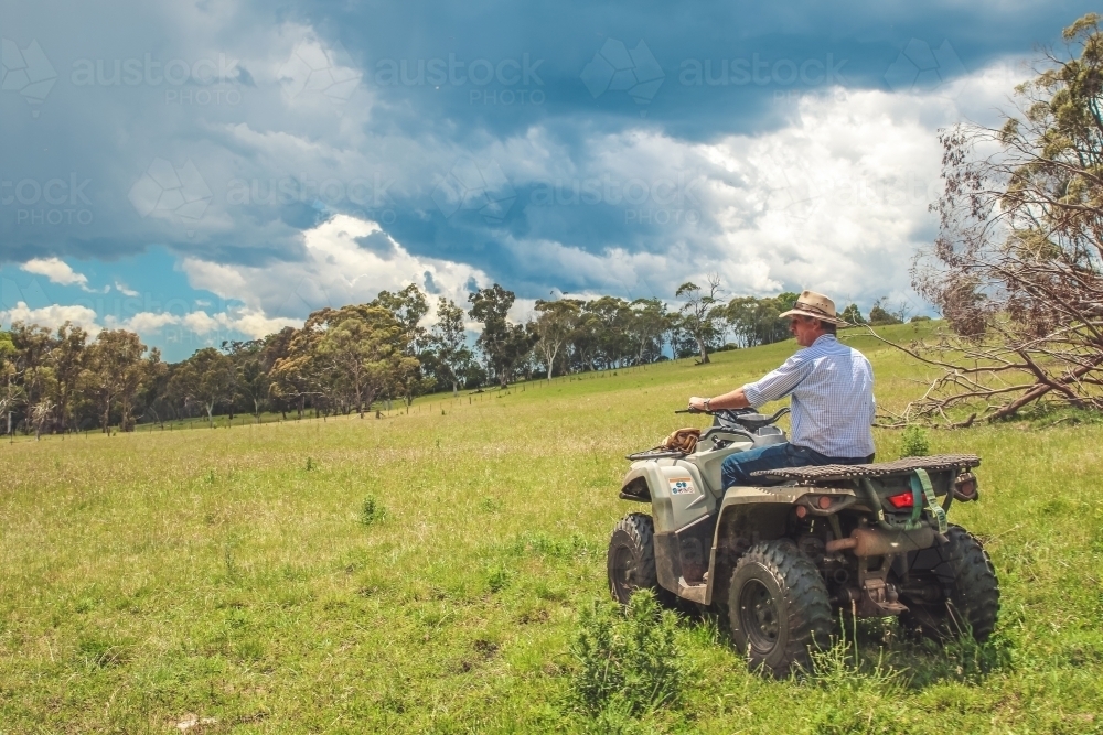 Male farmer sitting on stationary quad bike over looking paddock with dark storm clouds above - Australian Stock Image