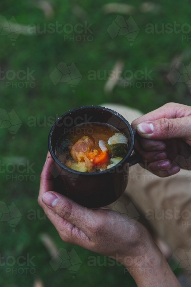 Male hands holding hot mug of soup, sitting on grass on cold afternoon - Australian Stock Image