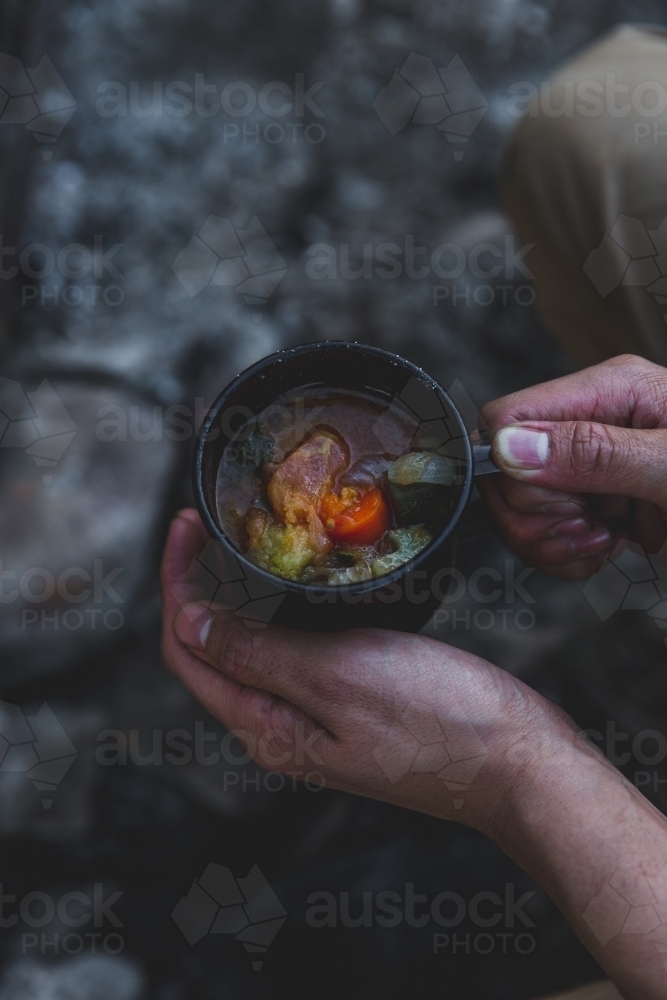Male hands holding warming mug of soup, sitting beside old campfire on cold evening - Australian Stock Image