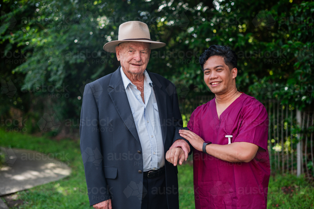 Male healthcare worker assisting an elderly man wearing a suit and wide brimmed hat outdoors - Australian Stock Image
