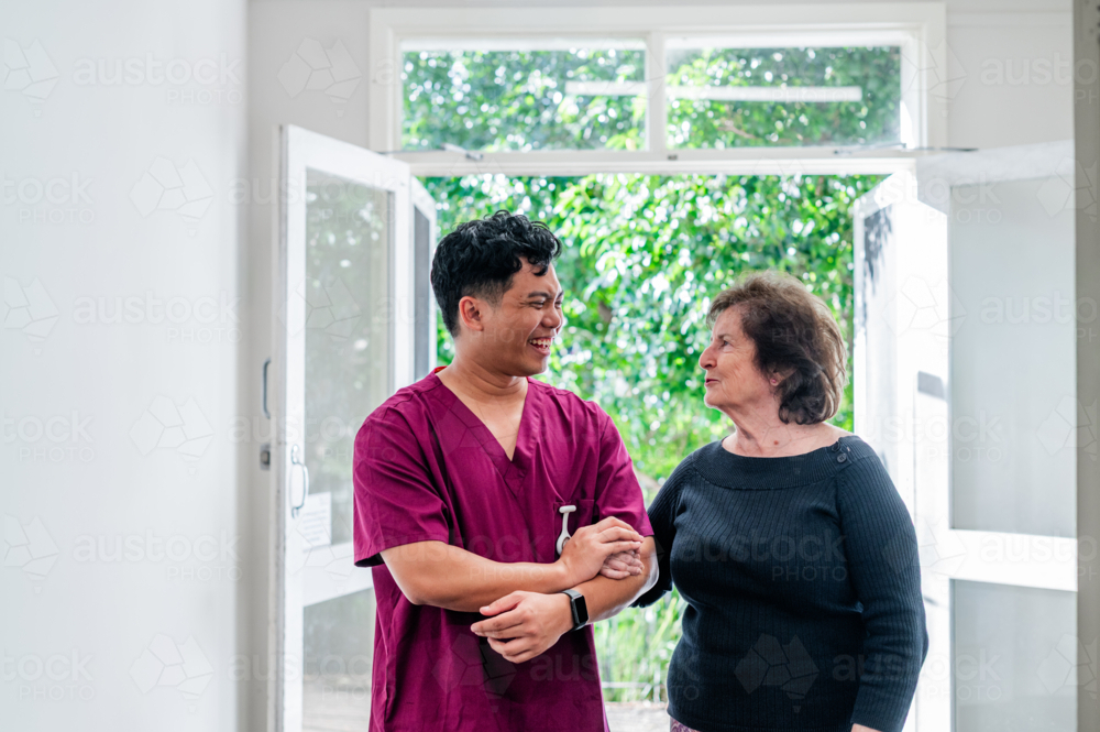 Male healthcare worker assisting an elderly woman walk indoors while having a conversation - Australian Stock Image
