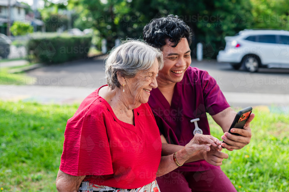 Male healthcare worker holding the phone for an elderly woman while seated on wooden bench - Australian Stock Image