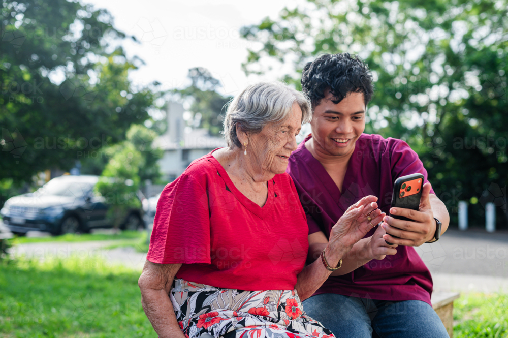 Male healthcare worker holding the phone for elderly woman seated on a bench outdoors on videocall - Australian Stock Image
