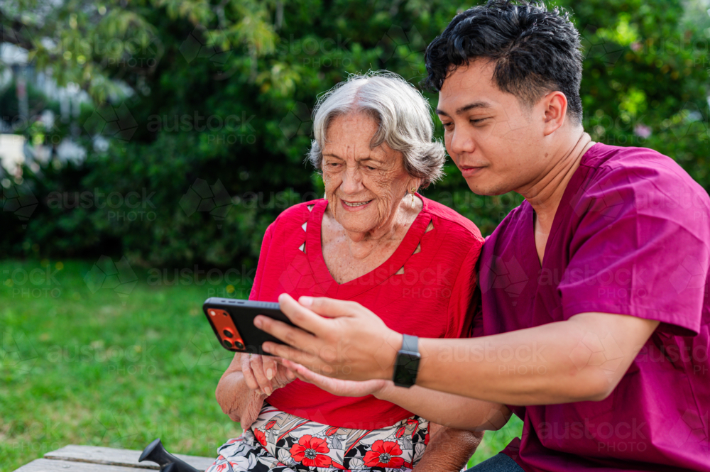 Male healthcare worker holding the phone for elderly woman seated on a bench outdoors on videocall - Australian Stock Image