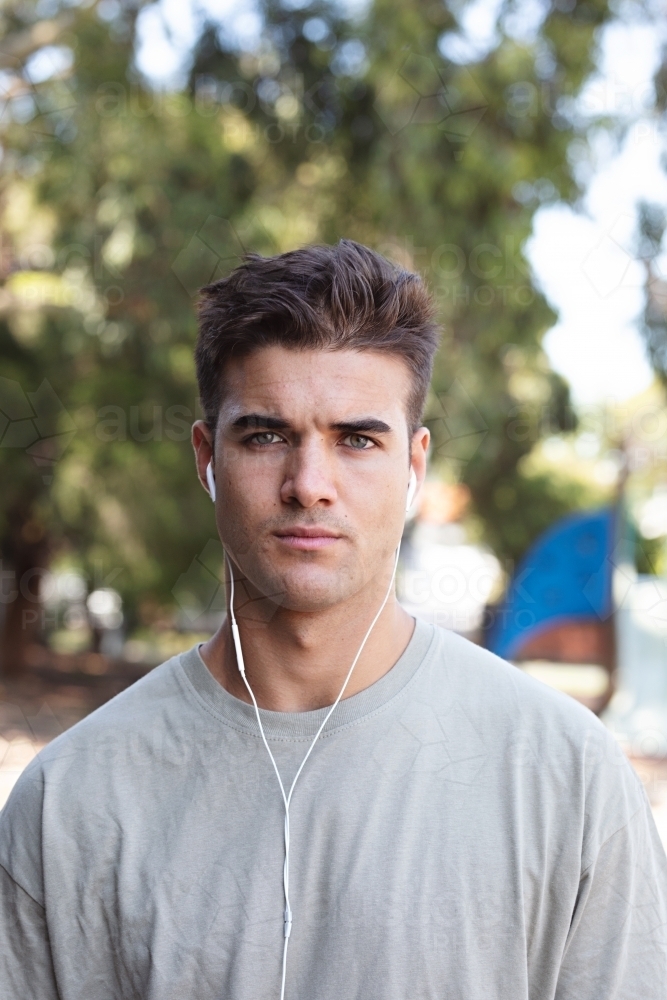Male in his twenties wearing earphones and listening to music outdoors in the local park - Australian Stock Image