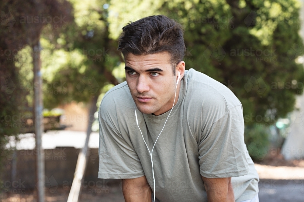 Male in his twenties wearing earphones and listening to music outdoors in the local park - Australian Stock Image