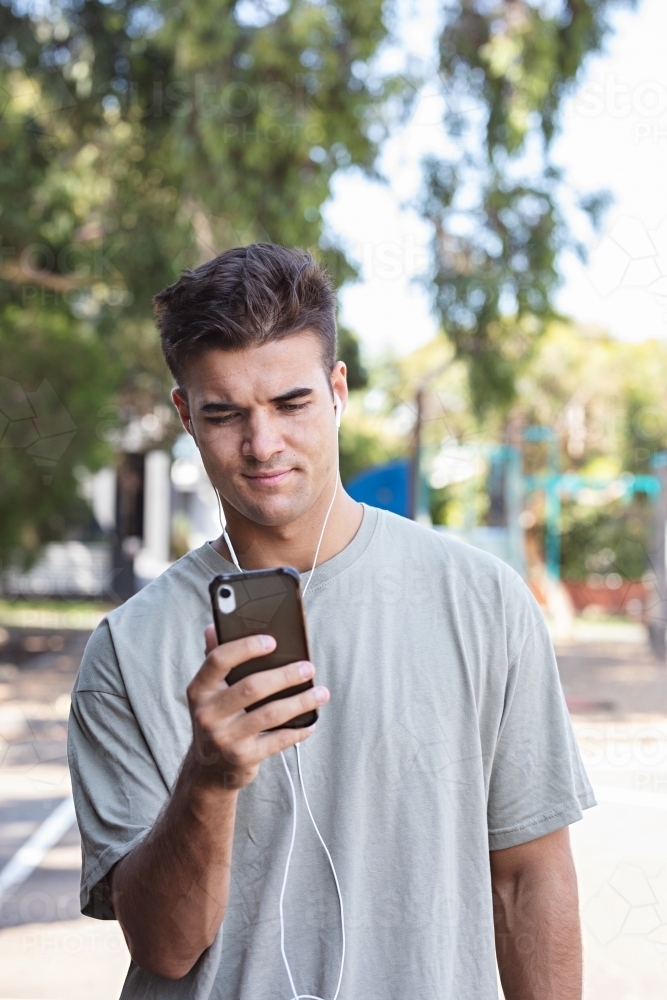Male in his twenties wearing earphones and listening to music outdoors in the local park - Australian Stock Image
