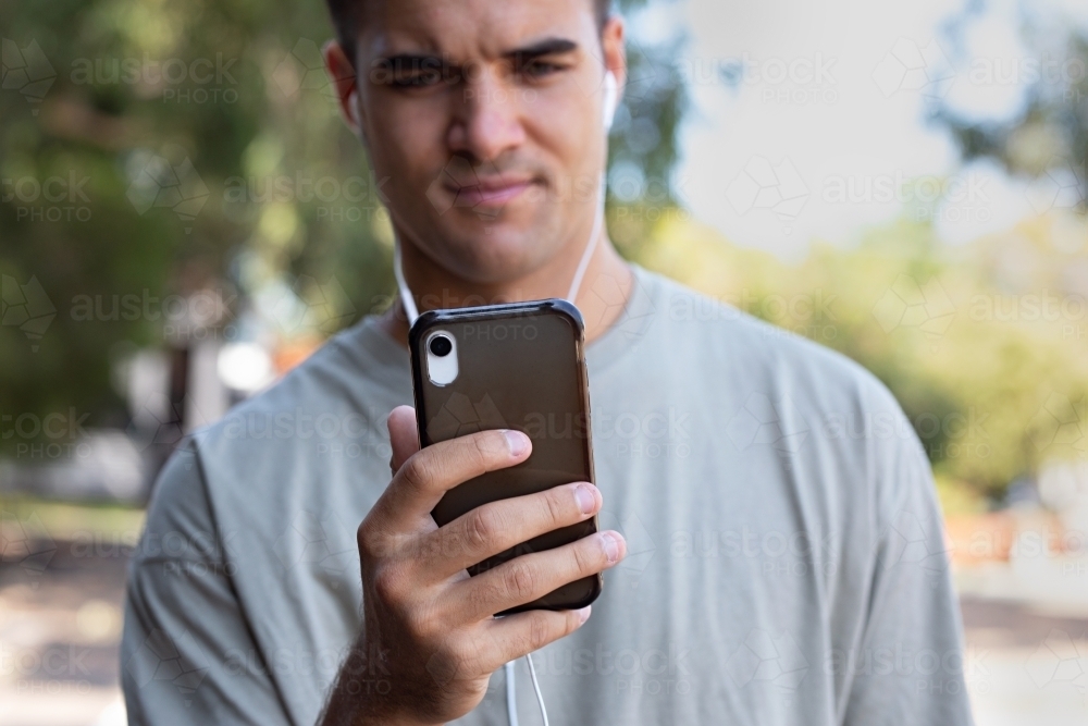 Male in his twenties wearing earphones and listening to music outdoors in the local park - Australian Stock Image