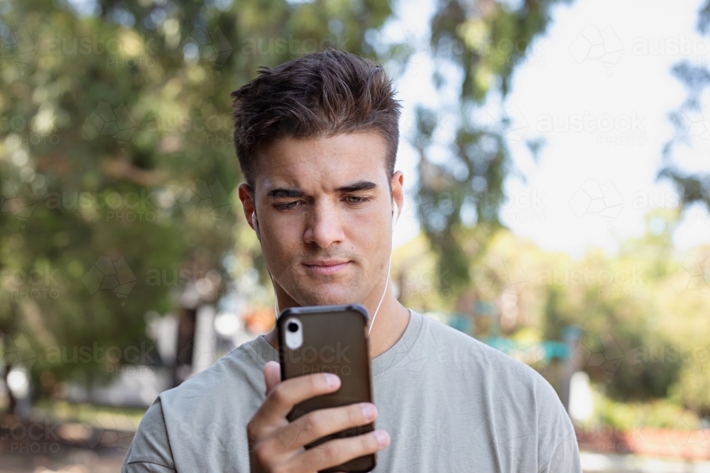 Male in his twenties wearing earphones and listening to music outdoors in the local park - Australian Stock Image