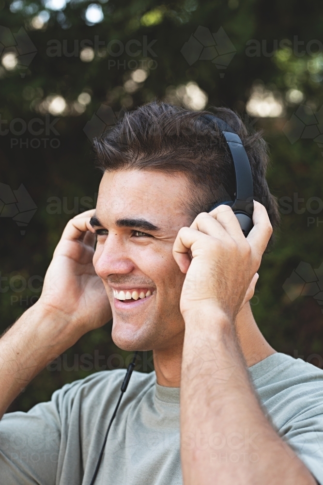 Male in his twenties wearing headphones and listening to music outdoors in the local park - Australian Stock Image