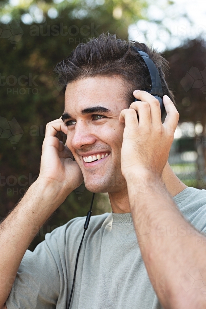 Male in his twenties wearing headphones and listening to music outdoors in the local park - Australian Stock Image
