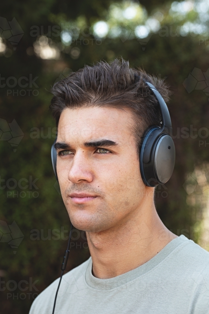 Male in his twenties wearing headphones and listening to music outdoors in the local park - Australian Stock Image