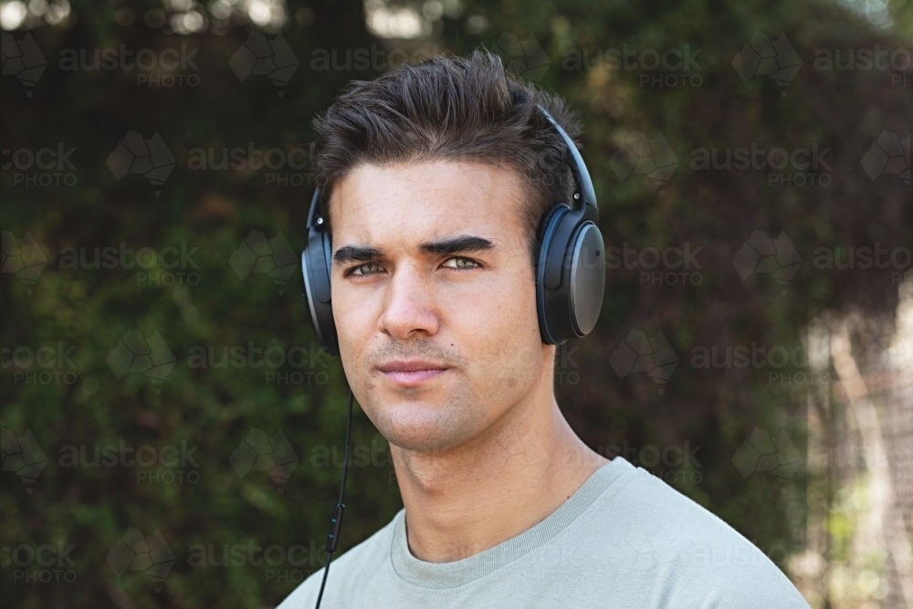 Male in his twenties wearing headphones and listening to music outdoors in the local park - Australian Stock Image