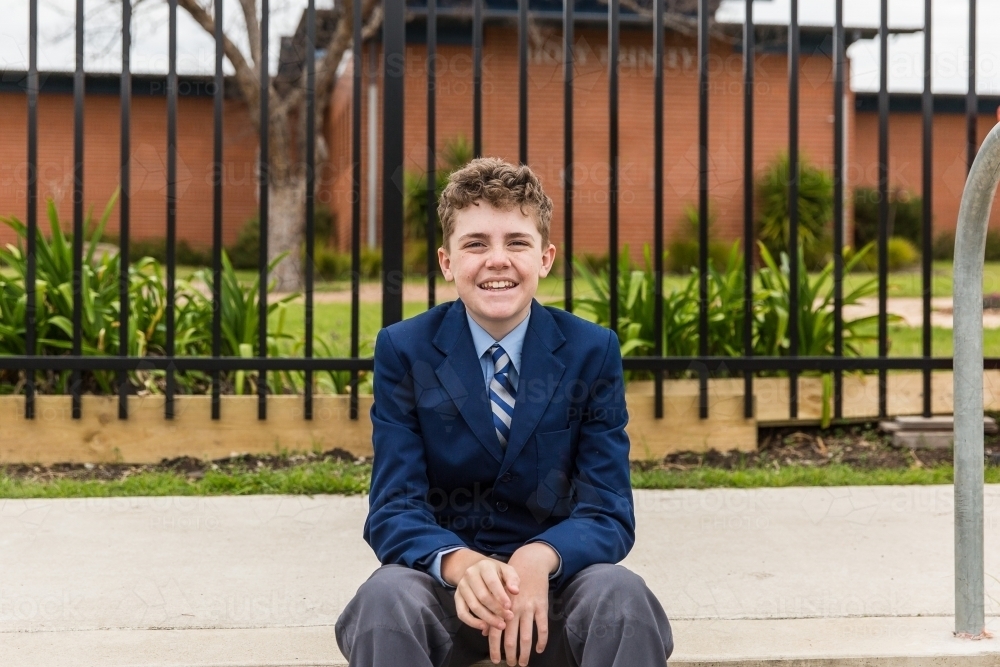 Male school child wearing uniform sitting on steps in front of school security fence smiling - Australian Stock Image
