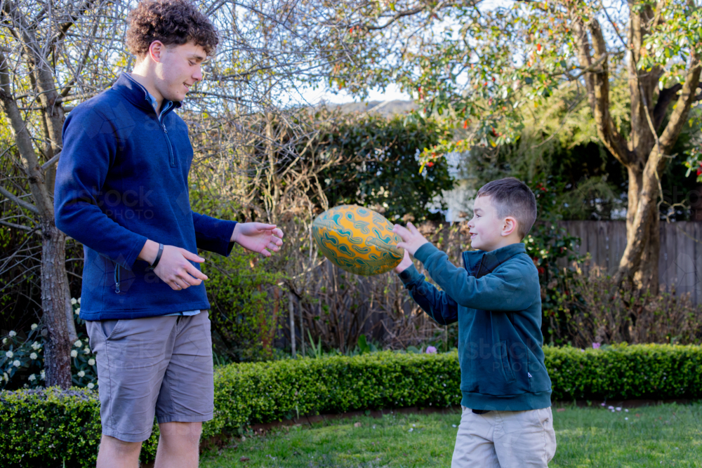 Male siblings passing the footy to each other in the backyard - Australian Stock Image