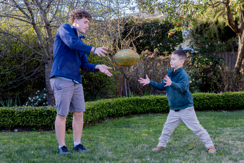 Male siblings passing the footy to each other in the backyard - Australian Stock Image