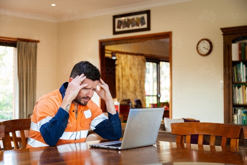 Male tradie in his 30s on laptop feeling overwhelmed and exhausted - Australian Stock Image