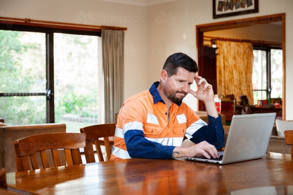 Male tradie in his 30s on laptop feeling overwhelmed and exhausted - Australian Stock Image