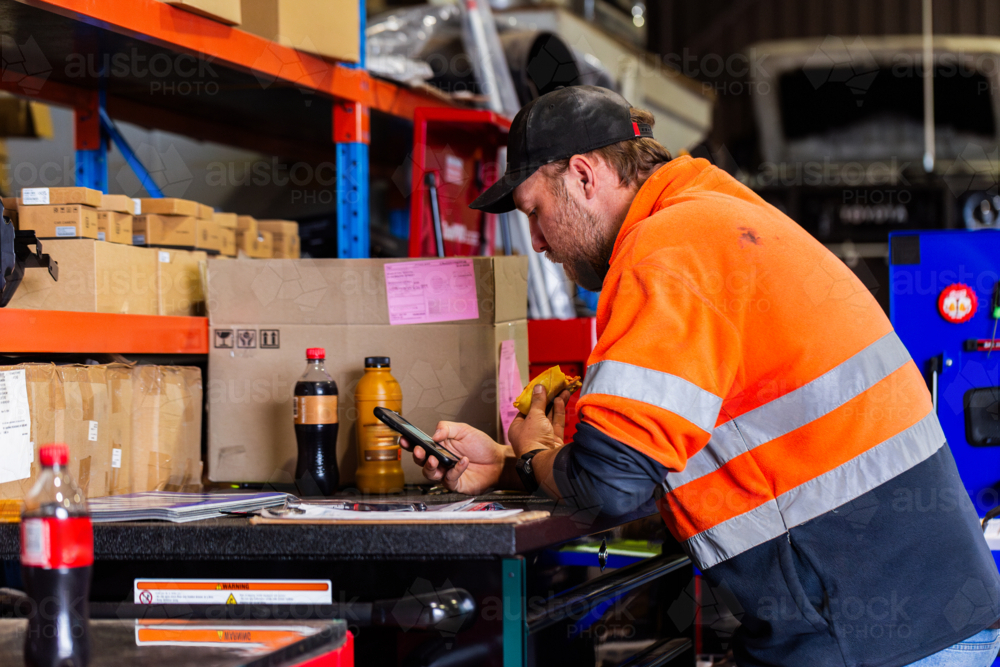 male worker on lunch break at industrial workplace using mobile phone eating meat pie - Australian Stock Image