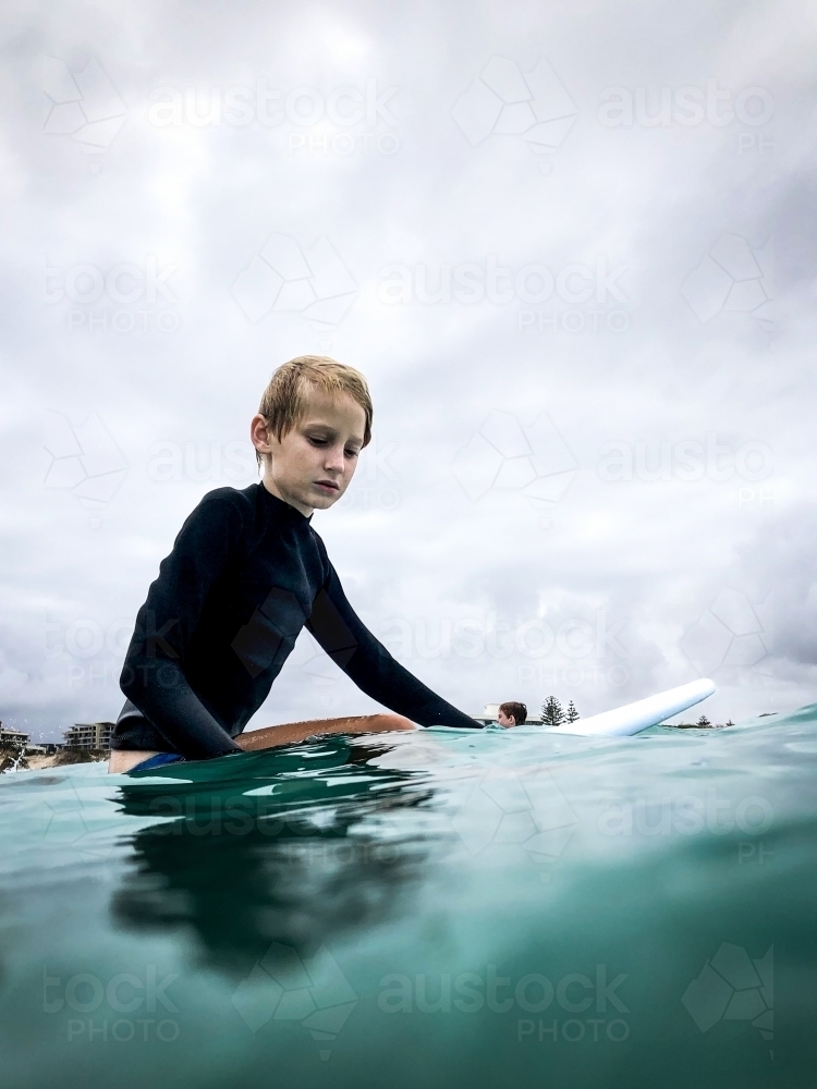 Male youth sitting on boogie board waiting for waves on overcast day - Australian Stock Image