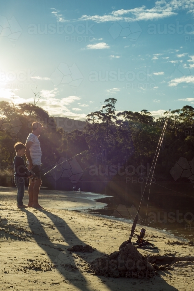 Man and boy fishing at river outlet with sun ray light - Australian Stock Image