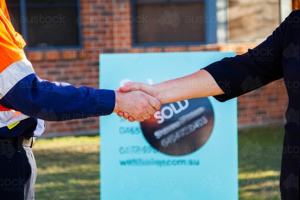 Man and businesswoman shake hands in front of house sold sign - Australian Stock Image