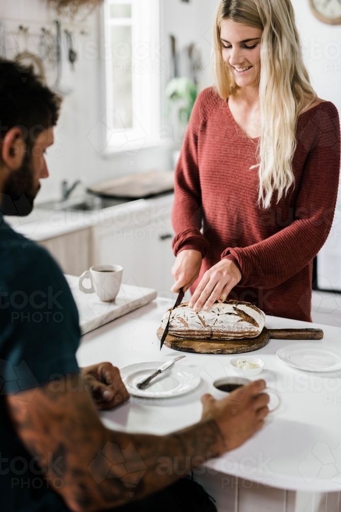 Man and woman at kitchen bench drinking coffee and slicing bread - Australian Stock Image