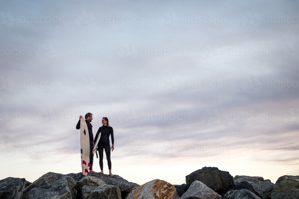 Man and Woman couple standing on coastal rocks holding surfboards looking at each other at sunset - Australian Stock Image