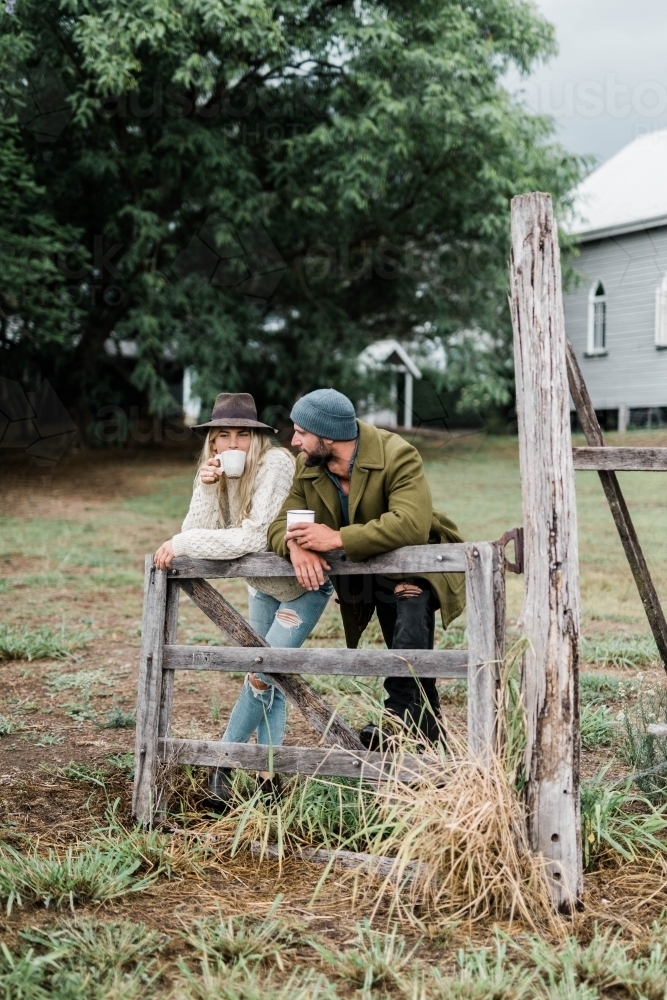 Man and woman leaning on wooden farm gate, drinking coffee - Australian Stock Image