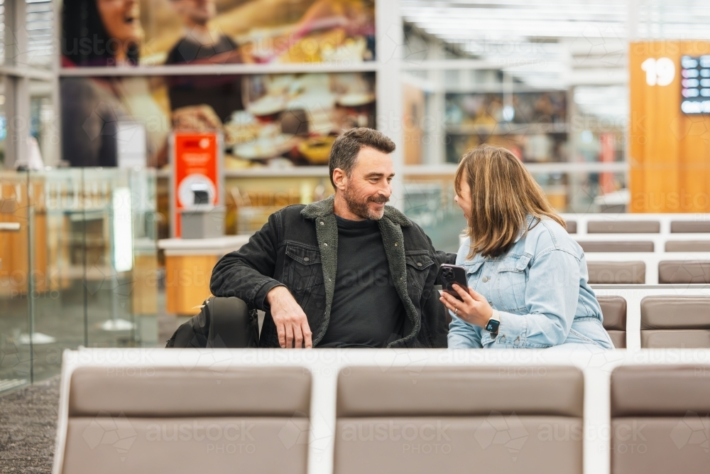 Man and woman watching phone together while waiting in airport departure lounge - Australian Stock Image