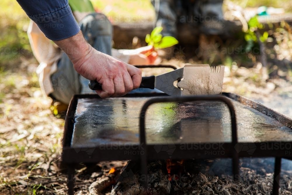 Image of Man cleaning hot plate for cooking sausages on campfire