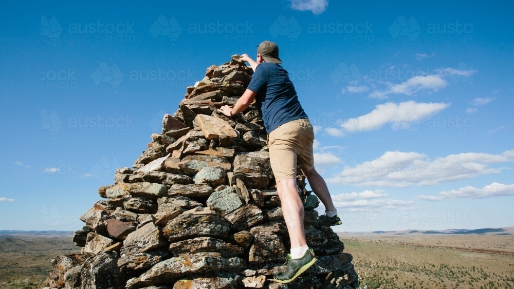 Man climbing a large rock cairn to balance a rock on the top - Australian Stock Image