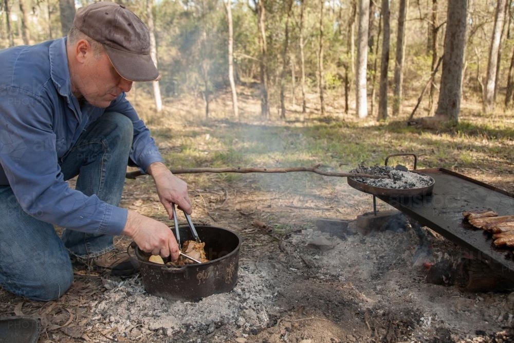 Man cooking meat in a camp oven on hot campfire coals - Australian Stock Image