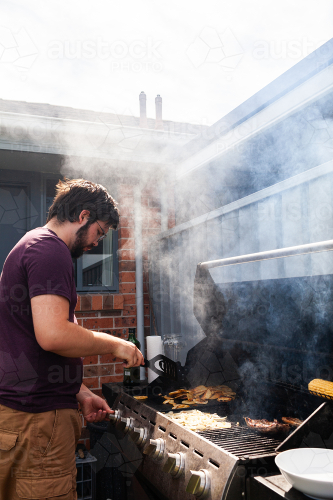 Man cooking potato chips on backyard barbecue in sunlight - Australian Stock Image