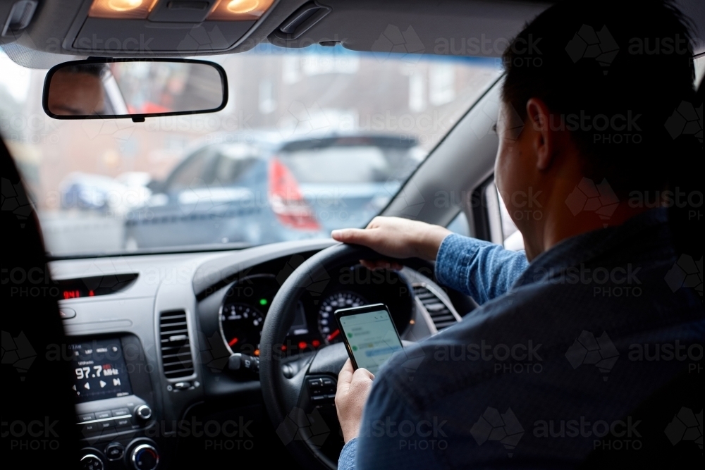 Man driving in his car looking down at mobile phone texting - Australian Stock Image